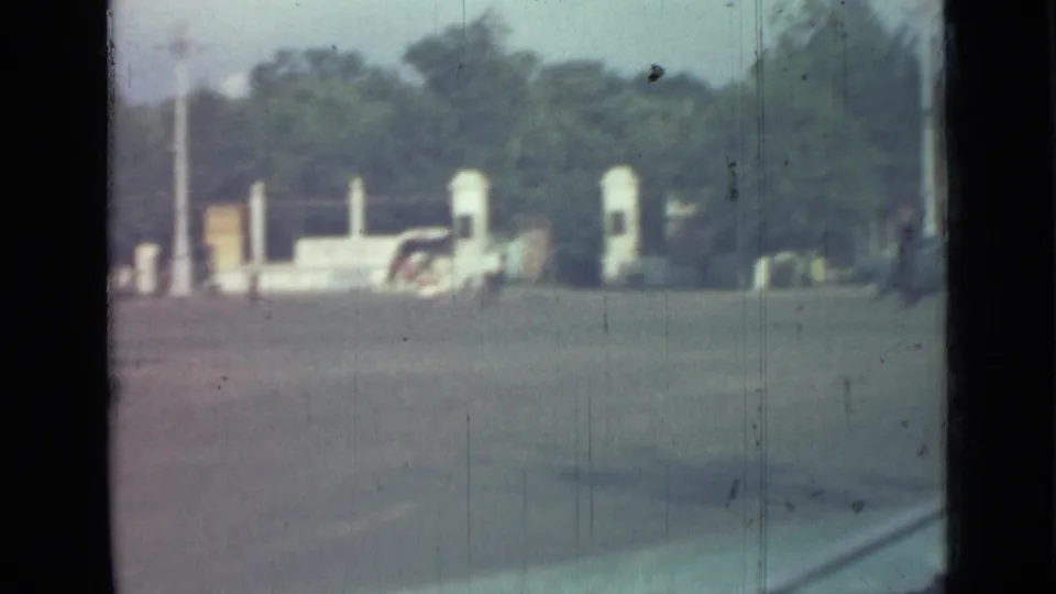 1967 home-movie frame with white gate or monument structures, a partly visible car, trees, and a wide paved foreground.