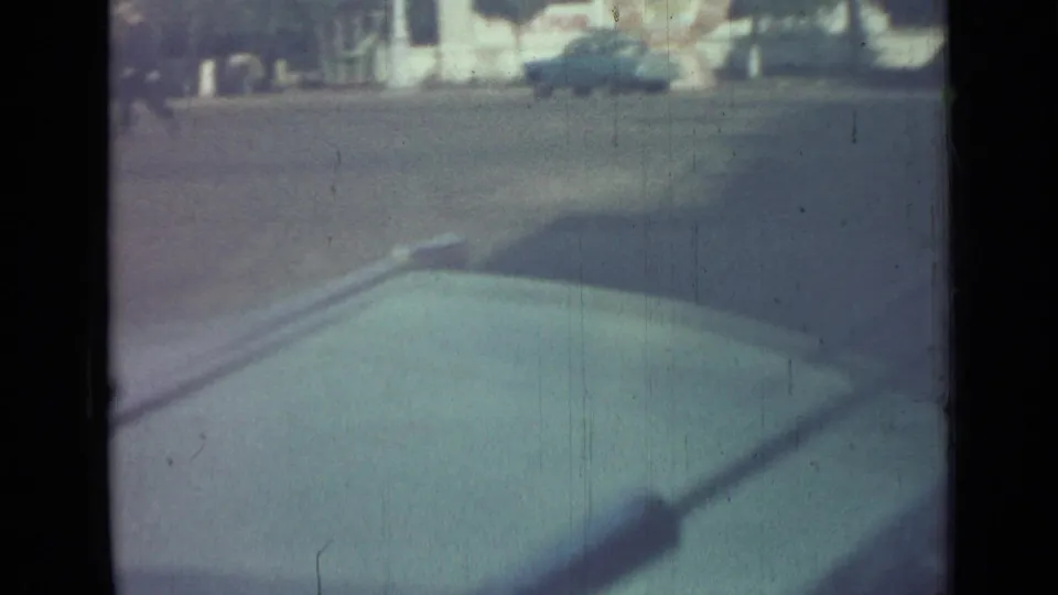 Hazy 8mm frame from the Saint Petersburg clip showing a blue car near the far curb and dark frame shadow over the pavement.