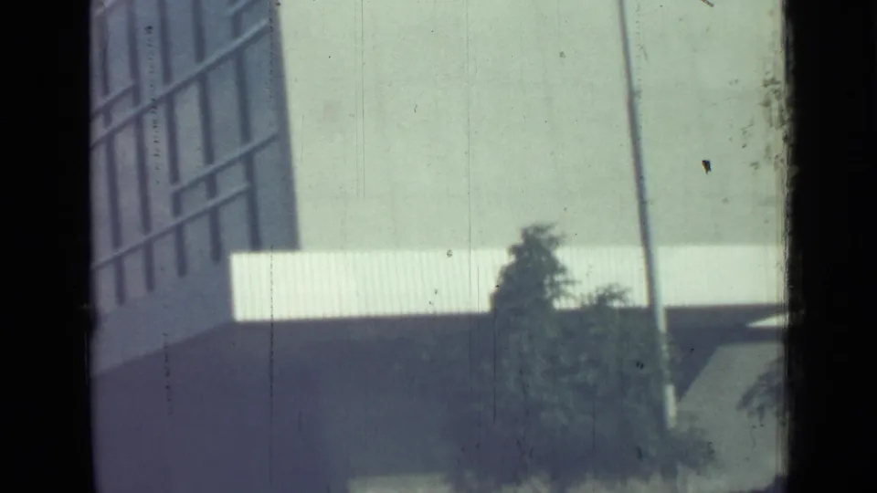 1967 Saint Petersburg home-movie frame with a blurred building wall, vertical pole, pale curb or railing, and dark planted greenery.