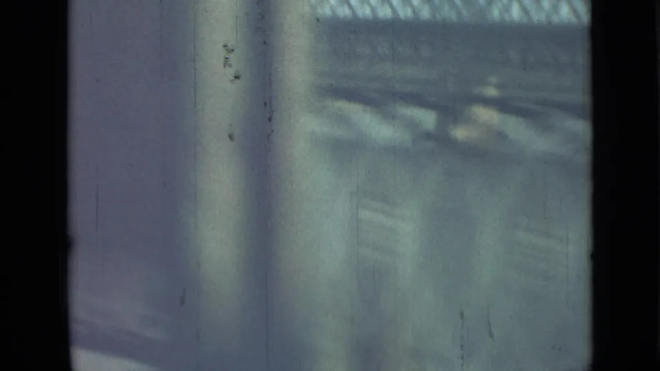 Muted 8mm frame from a moving vehicle, showing a vertical window obstruction and an indistinct railing or barrier outside.