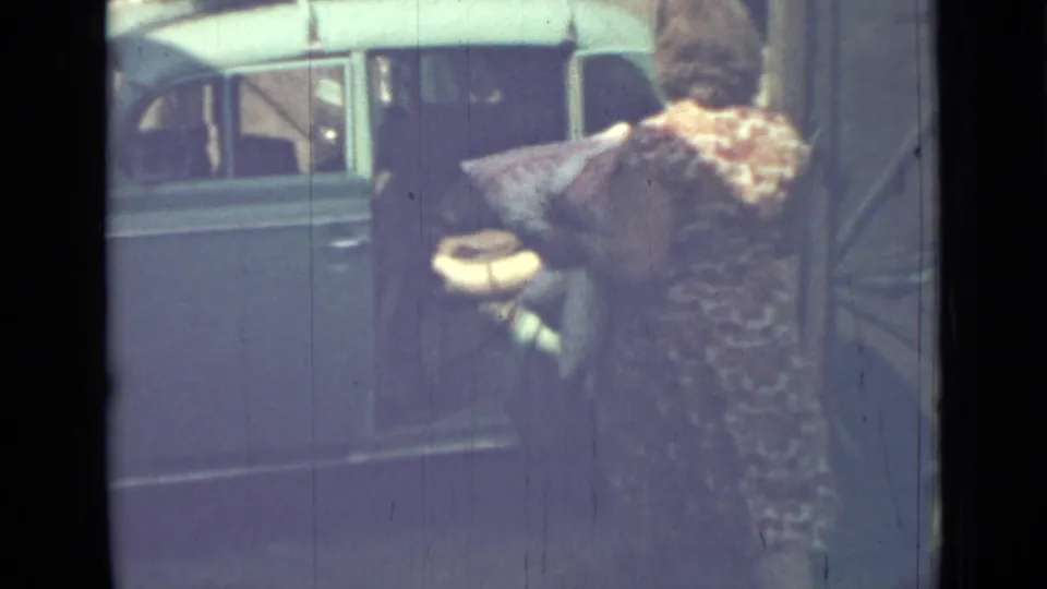The woman stands with her back to the camera, holding soft items beside the open blue van in the 1967 home-movie scene.