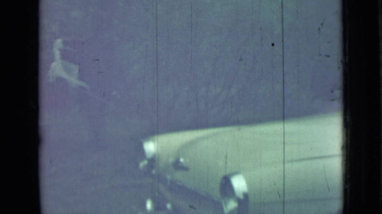 Front portion of a pale vintage car seen through dim glass and film scratches.