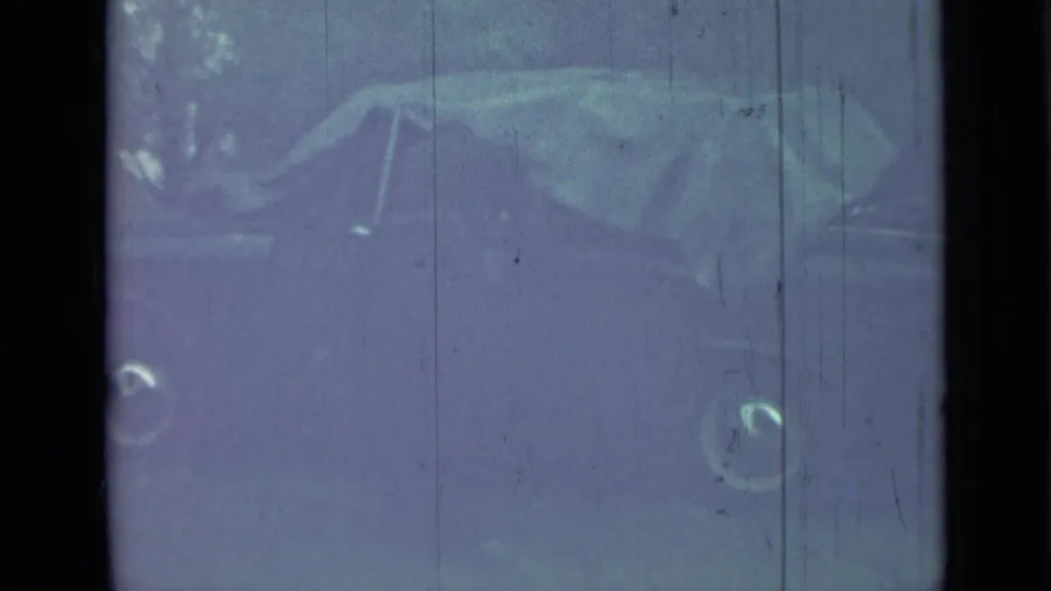 Obscured front or side-front view of the parked sedan, softened by exposure, reflections, and 8mm film wear.