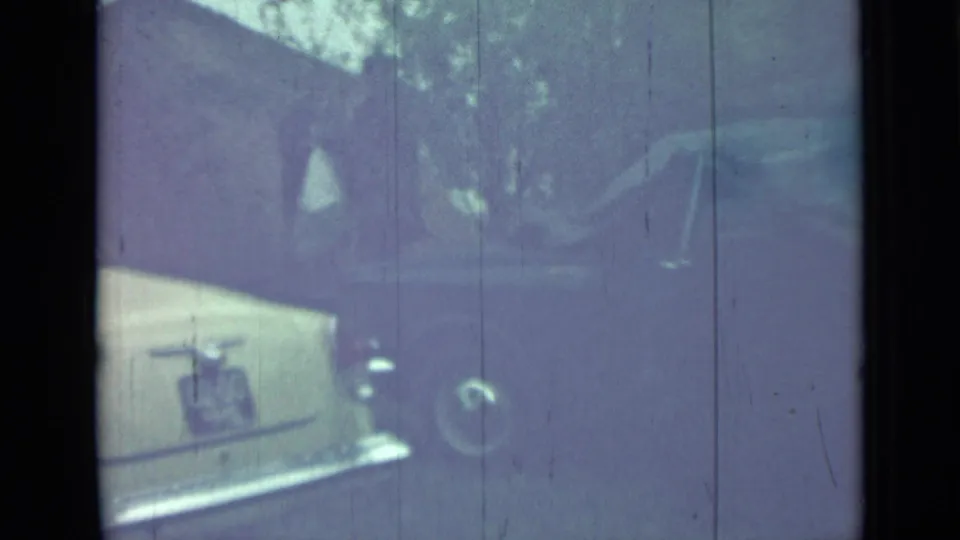 Partial rear-side view of the pale sedan in Saint Petersburg, with trees and driveway surroundings in 1967 8mm footage.