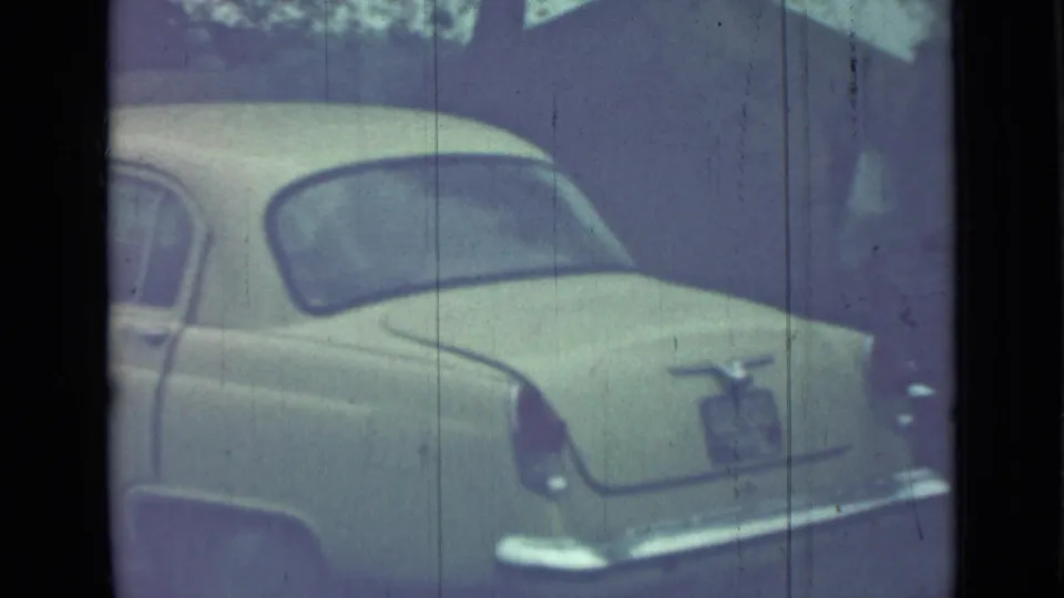 Rear view of the pale 1960s-style sedan, showing trunk, rear window, chrome bumper, and plate area in 8mm footage.