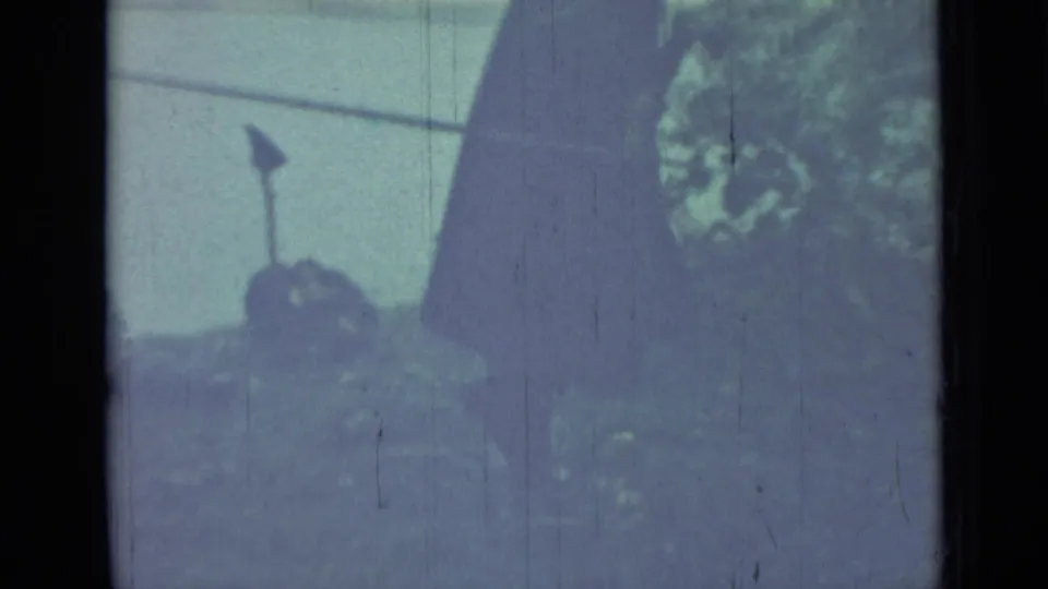1967 Saint Petersburg 8mm frame with part of a dark-clothed figure beside grass, trees, and outdoor equipment near water.