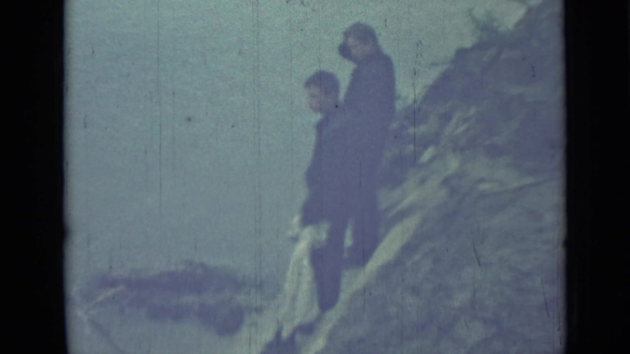 Two dark figures stand on a sloped hillside against a pale sky.
