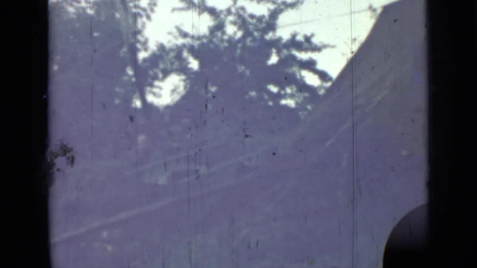 1967 8mm frame with trees, pale sky, and a curved dark edge that may be part of the abandoned vehicle described in the clip metadata.