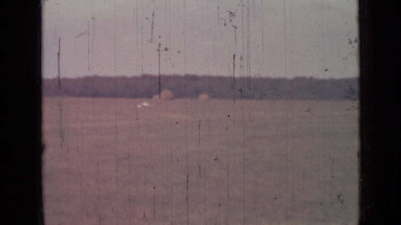 Open field and distant tree line seen through dark vertical framing on scratched 8mm film.