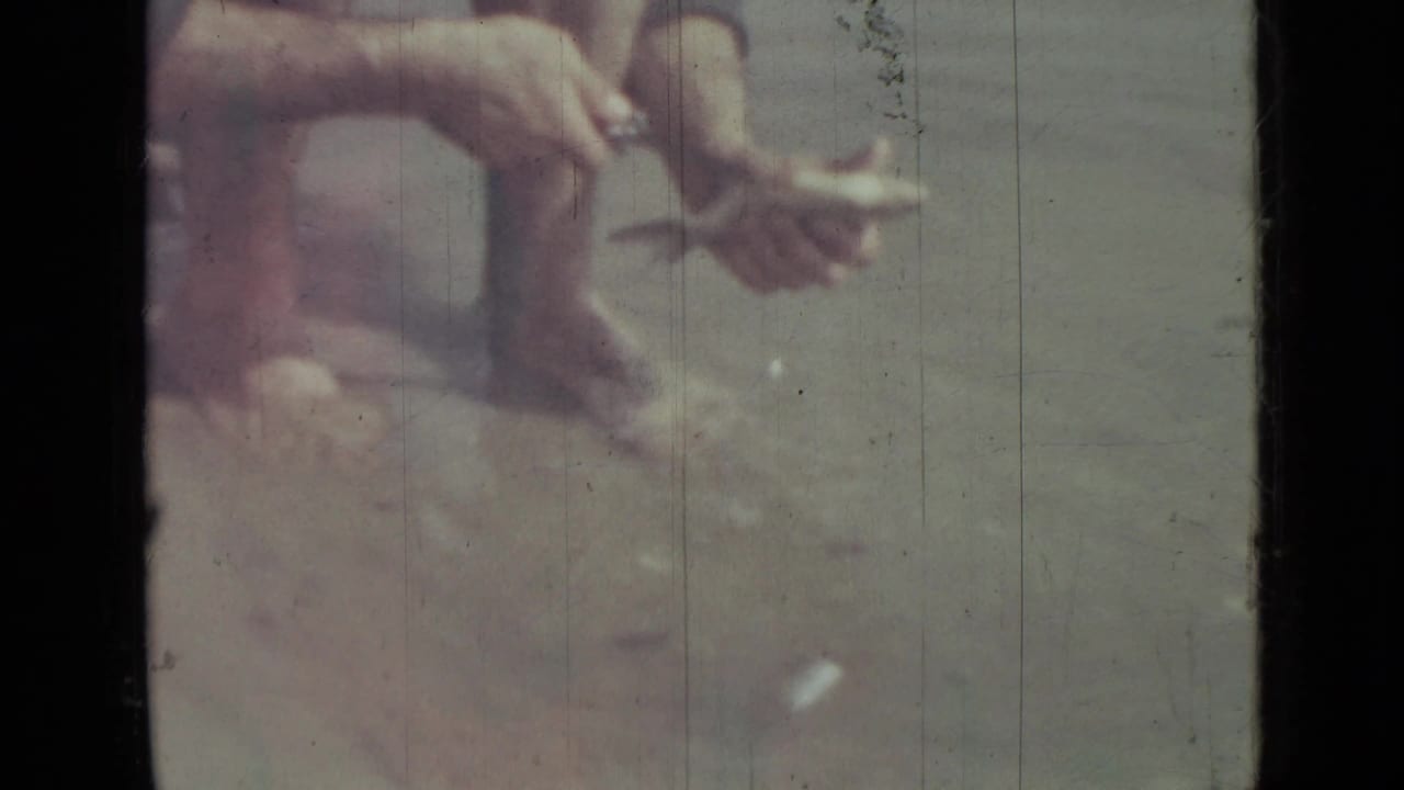 Barefoot man squatting near water while holding a fish and knife.