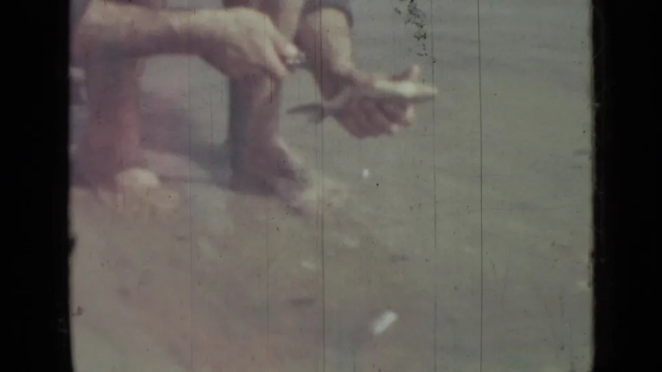 1967 8mm home-movie frame from Saint Petersburg showing a barefoot man crouched at the water's edge, working on a fish with a small knife.