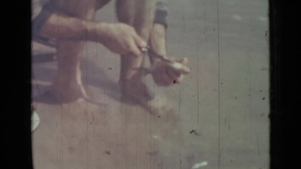Close 8mm frame shows the man's hands cutting into a fish while he squats barefoot by the water in the Saint Petersburg home-movie footage.