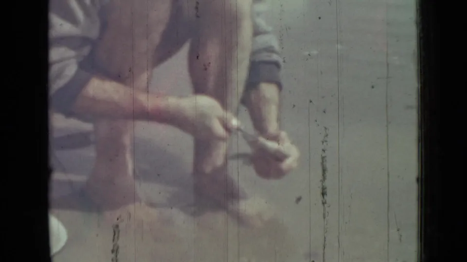 The home-movie frame shows the man crouched at the shoreline, holding the fish while a white bowl rests near his knee.