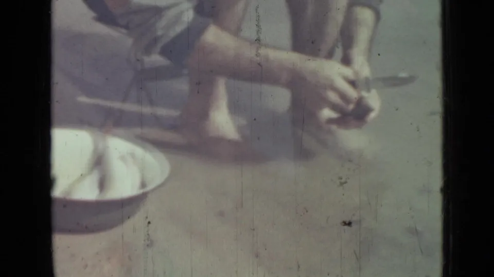 Close 1967 8mm frame shows the man continuing to gut the fish, with a white bowl placed beside him on the wet shoreline.