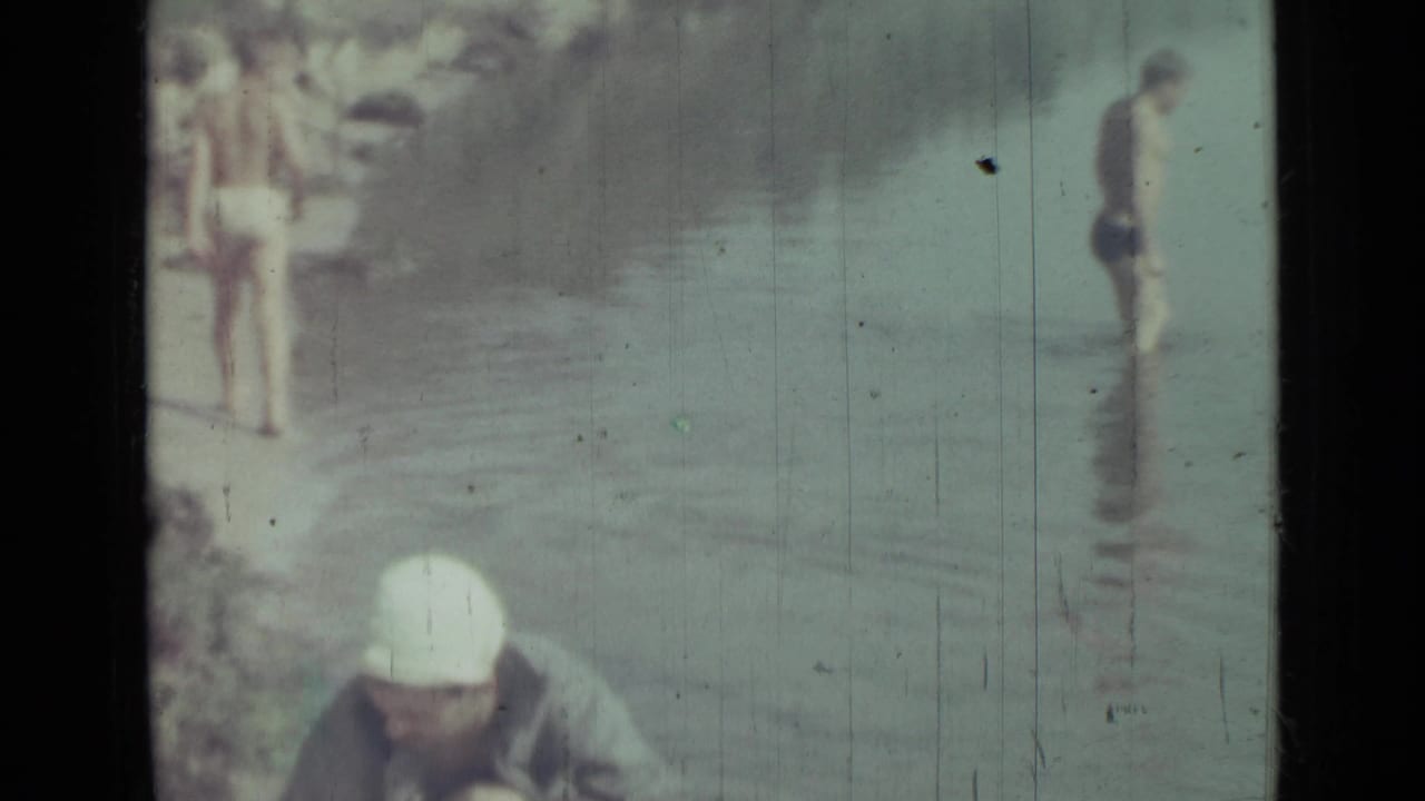 Several figures in swimwear stand and wade in shallow water on faded 8mm film.