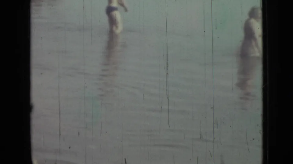 Faded 8mm frame from the Saint Petersburg clip with young figures standing in shallow water and heavy vertical film wear.