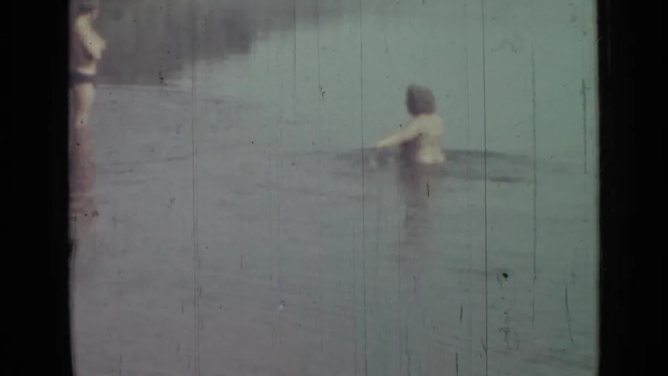1967 home-movie frame showing a young person chest-deep in water, with another partial figure at the left edge and faded 8mm texture.