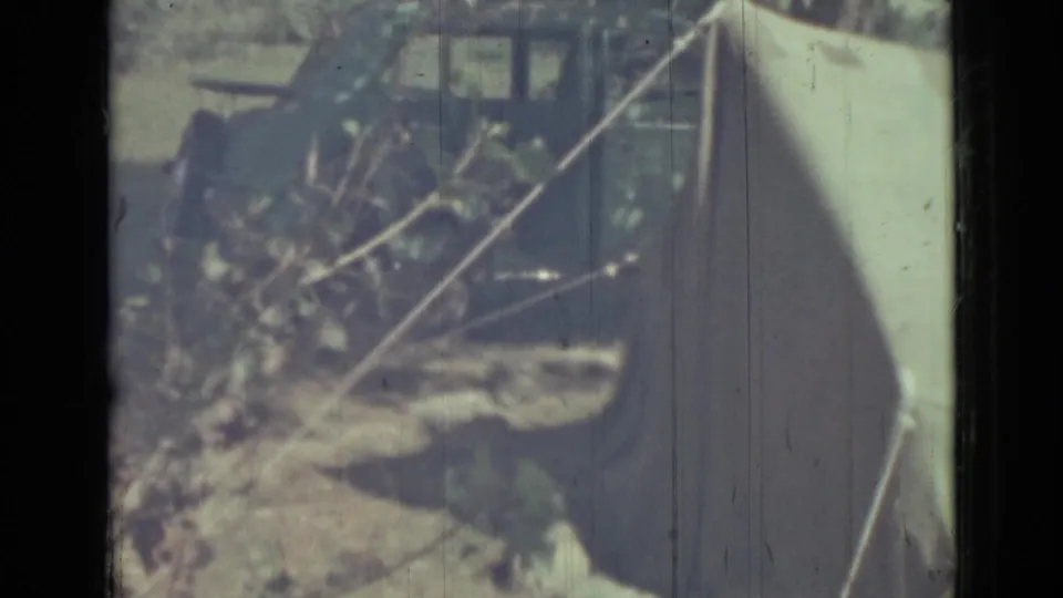 Handheld 8mm view of the tent edge, branches, and guy line, with a dark rectangular vehicle-like or structure-like form in the background.