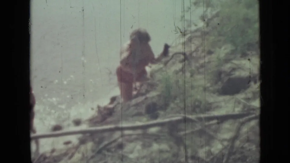 Distant woman in swimwear on rocky shoreline beside pale water, framed by foliage in a 1967 Saint Petersburg home-movie context.