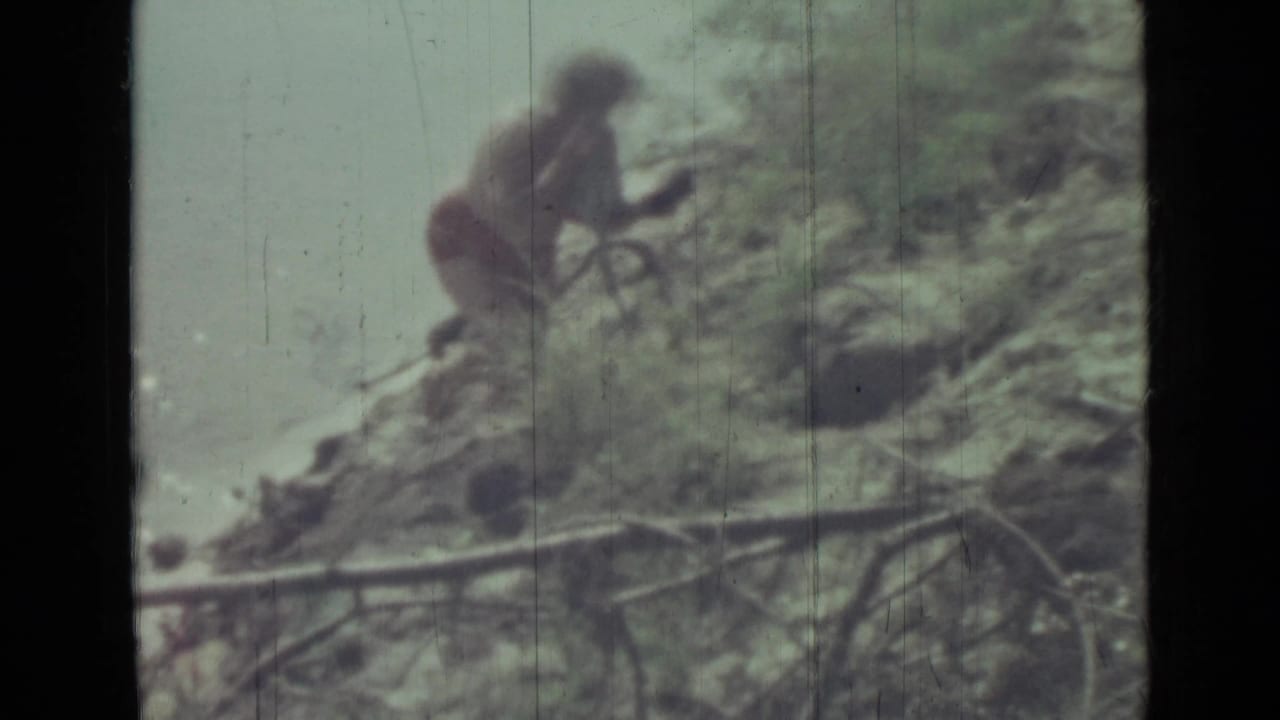 Woman in a red bikini bending or stepping near rocks and foliage outdoors.