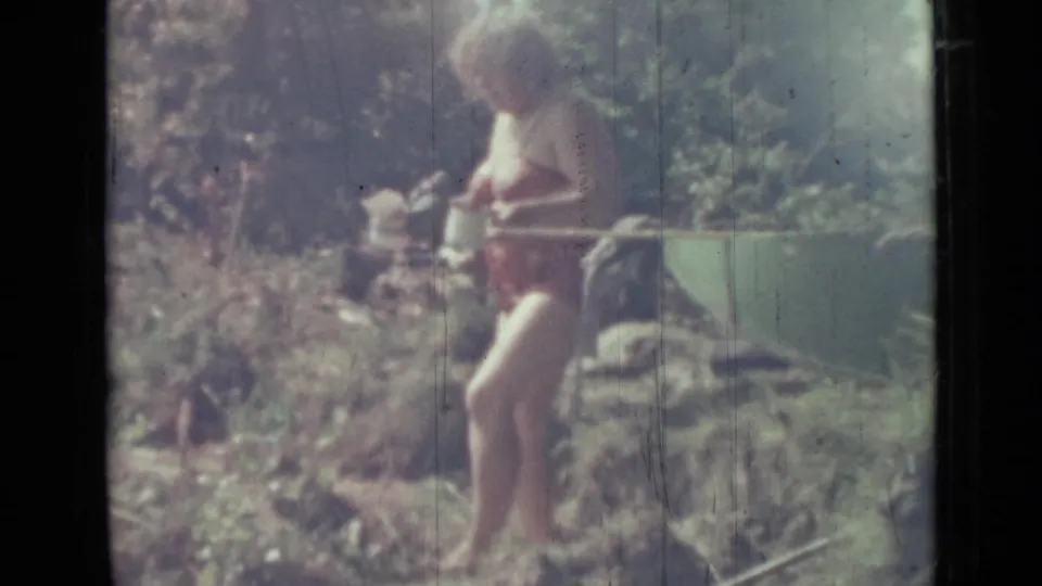 1967 Saint Petersburg home-movie frame of a woman in red swimwear walking through brush with a white container in hand.