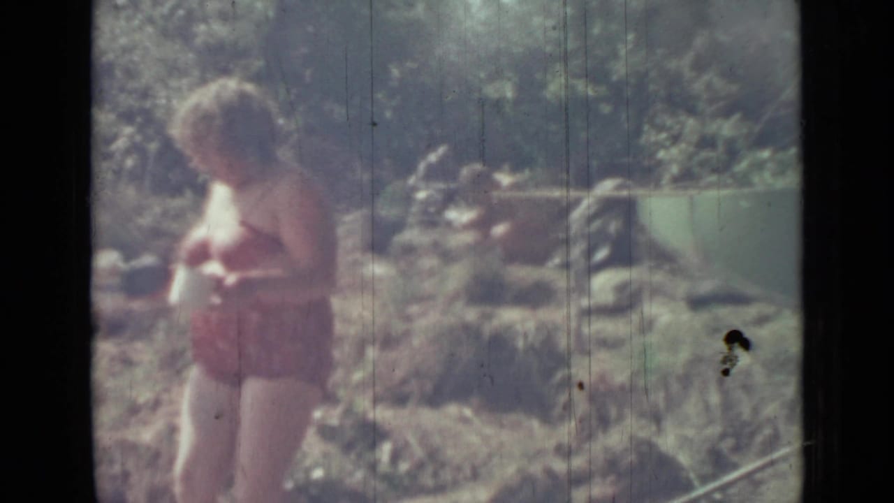 Older woman in a red swimsuit holding a white mug near a rocky, grassy riverside.