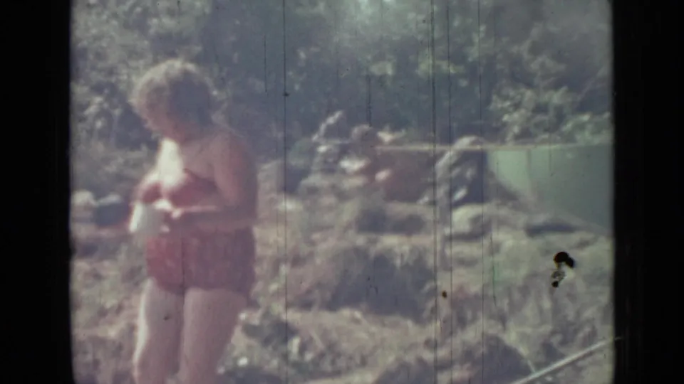 1967 Saint Petersburg home-movie frame of an older woman in a red swimsuit holding a mug beside rocks and greenery near water.