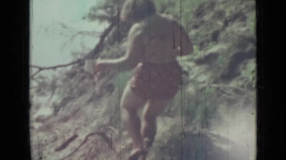 1967 home-movie frame from Saint Petersburg showing the woman from behind near branches, stones, and sunlit riverside vegetation.