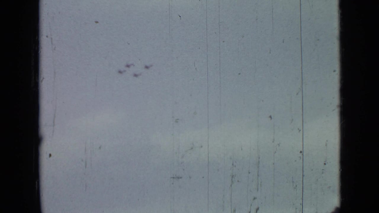 Distant planes crossing a pale scratched sky in an 8mm film frame.