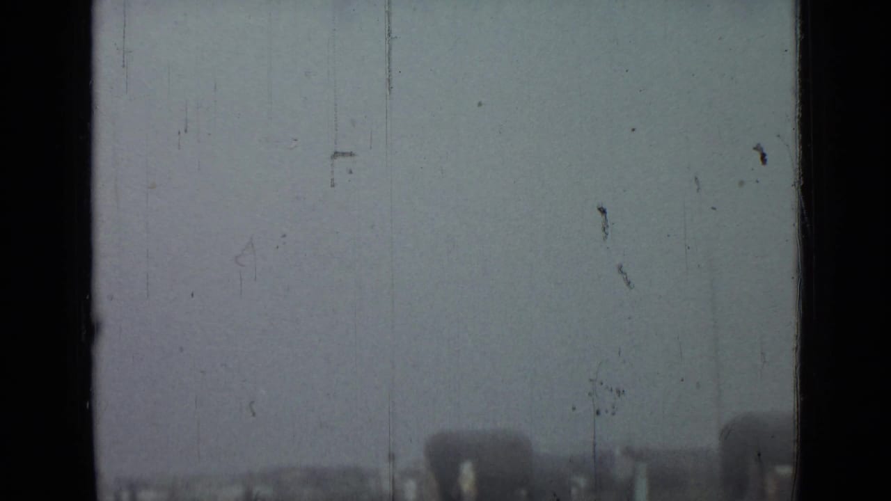 Hazy 1967 Saint Petersburg home-movie frame with a grassy field, distant arched structure, scratches, and soft exposure.