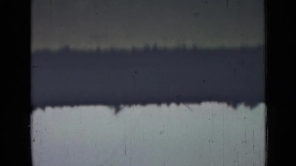 Faded 8mm frame with a dark exposure band, grassy lower area, hazy sky, scratches, and indistinct distant shapes.