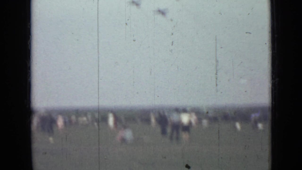 Distant people stand in a field while small aircraft fly above the horizon.