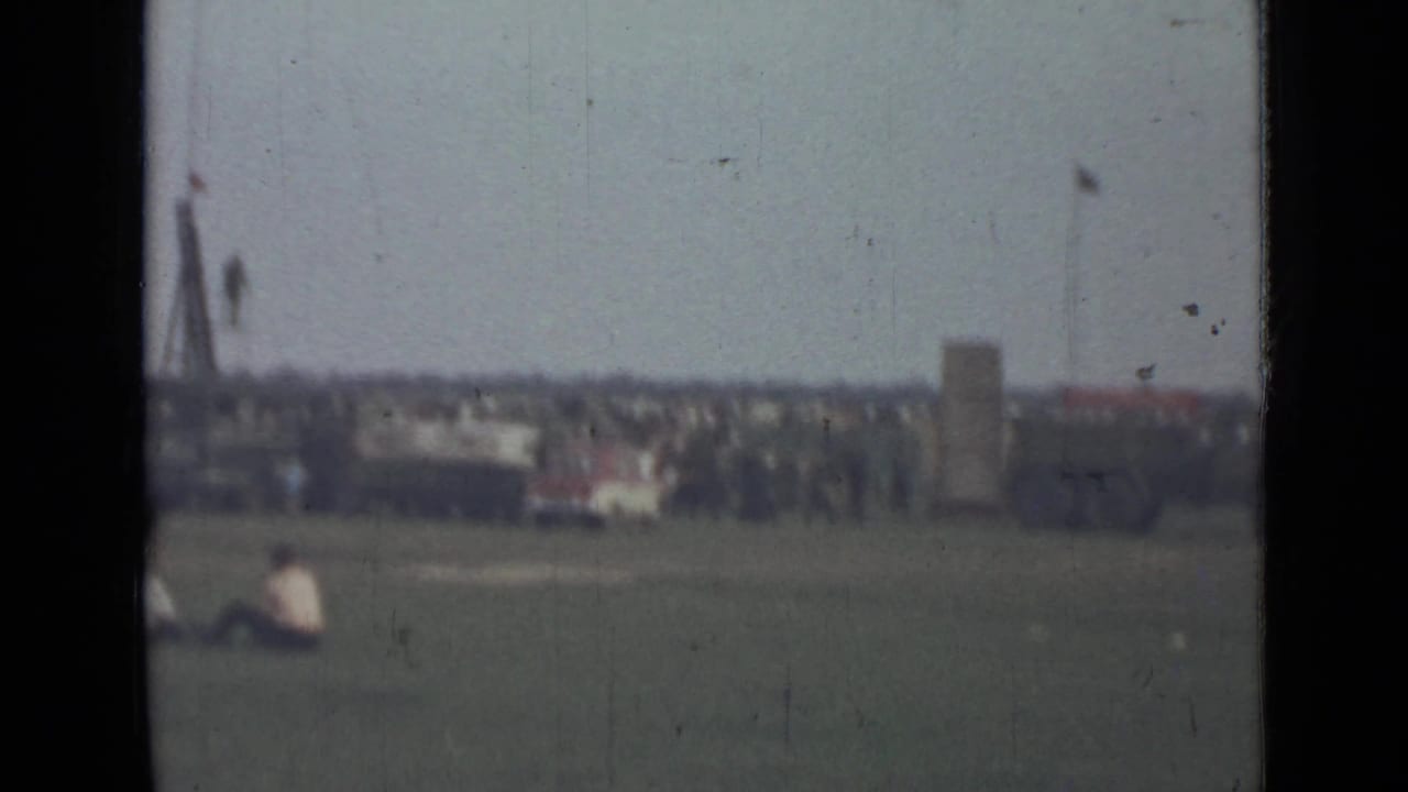 1967 8mm view near Saint Petersburg showing a wide field, seated spectators, and a distant crowd near a vehicle and tall poles.