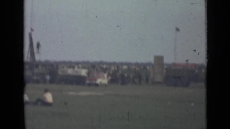 1967 8mm view near Saint Petersburg showing a wide field, seated spectators, and a distant crowd near a vehicle and tall poles.