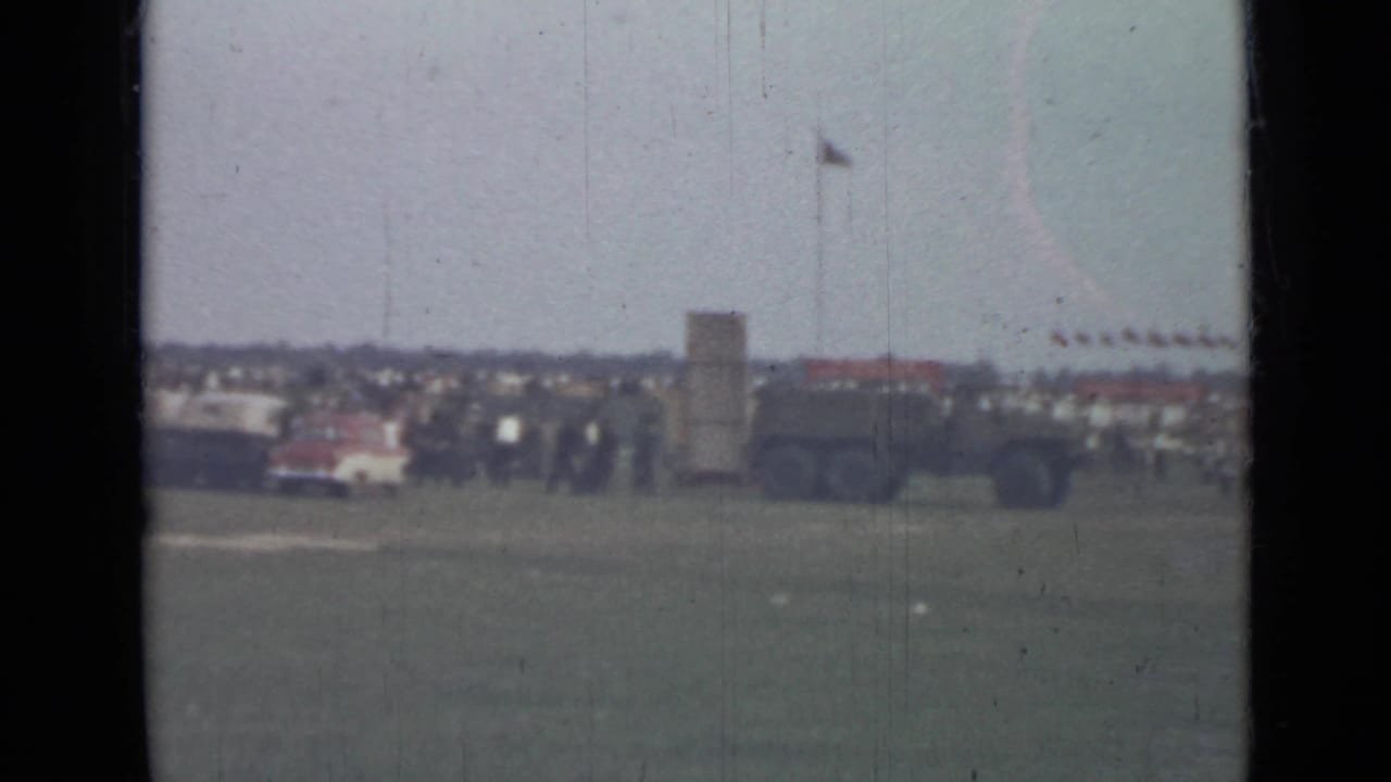 Distant Saint Petersburg field scene with a crowd, a military-style vehicle, tall antenna or pole, and low buildings beyond the grass.