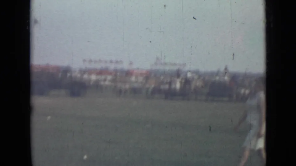 Soft 8mm frame of a 1967 Saint Petersburg field gathering, with distant spectators and a woman partly entering the foreground.