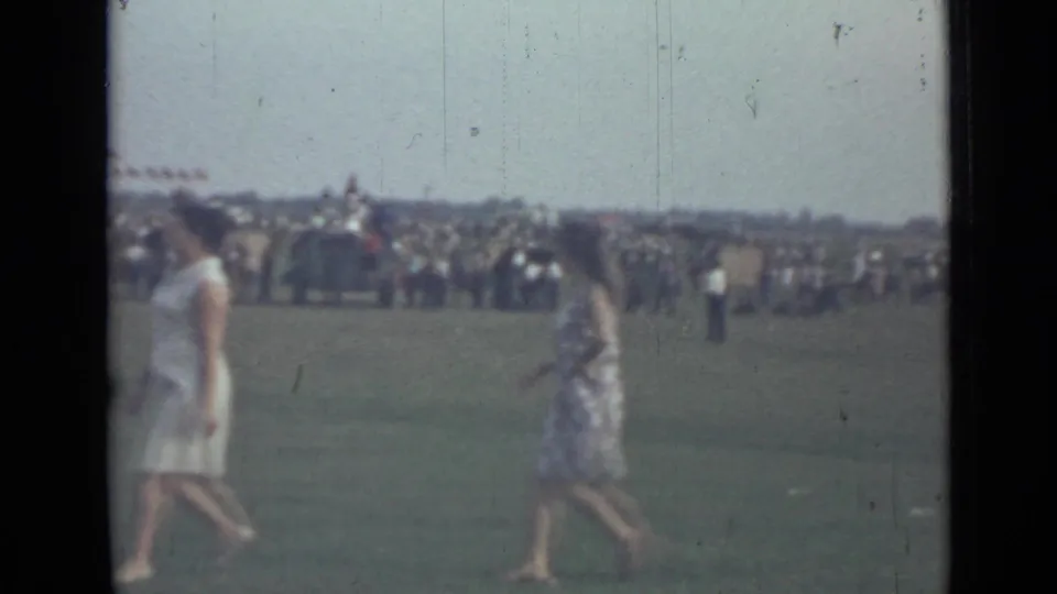 1967 home-movie frame near Saint Petersburg with two women crossing the grass before a distant crowd, flags, and vehicles.