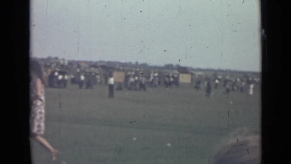 1967 Saint Petersburg home-movie frame showing a distant crowd on a grassy field and a woman partly visible at the left edge.