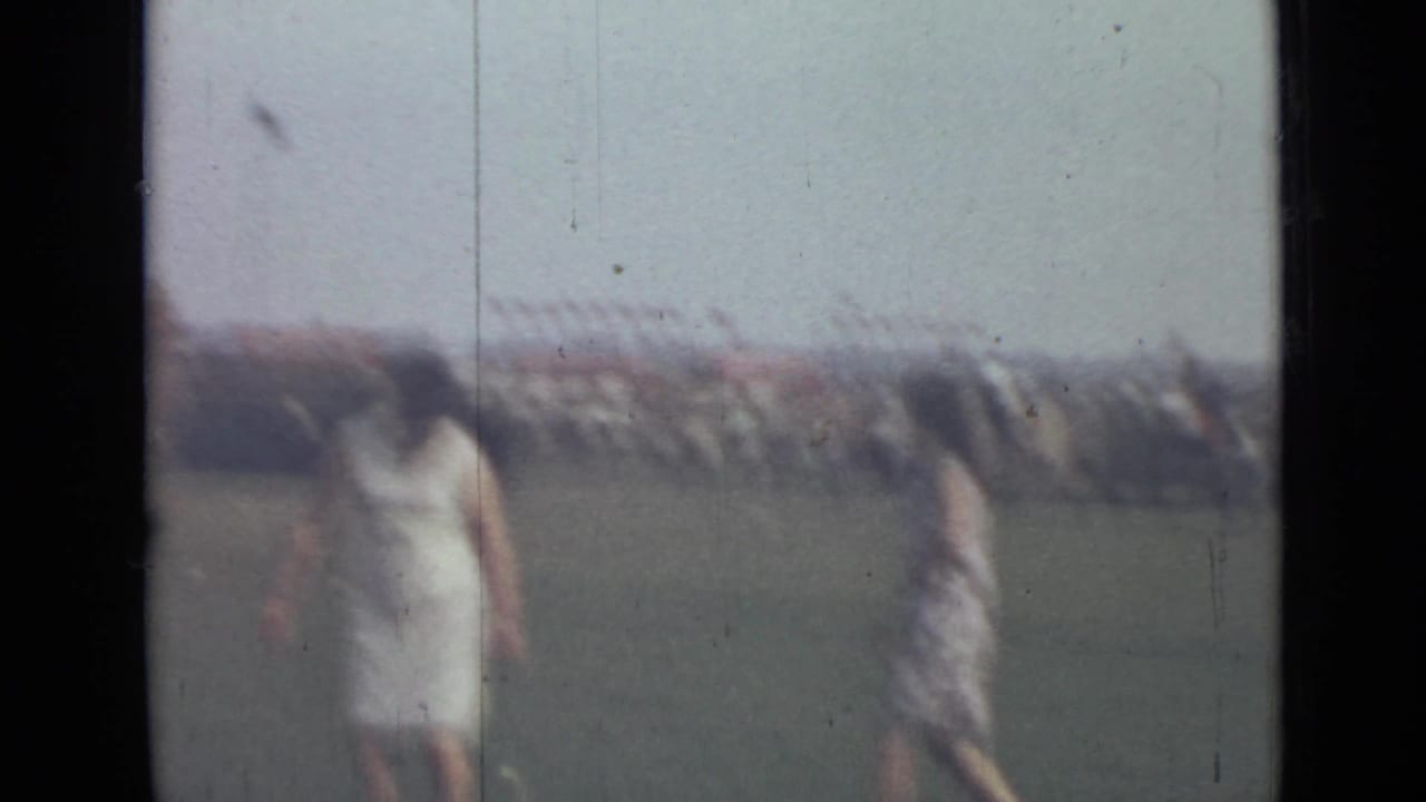 1967 Saint Petersburg home-movie frame with two women walking across a grassy field before a distant public gathering.