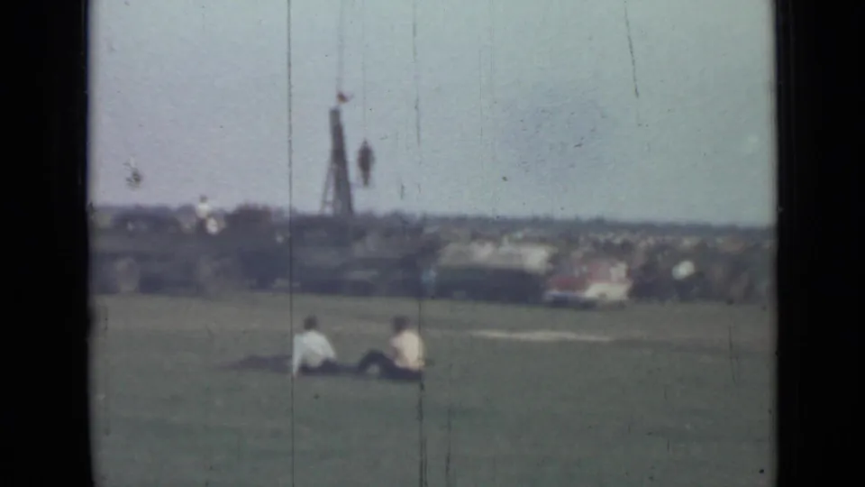 1967 Saint Petersburg home-movie frame showing seated figures on a field with distant buildings and a tall structure beyond.