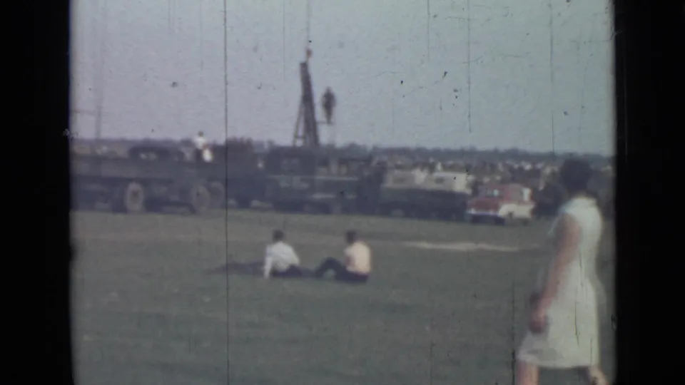 1967 Saint Petersburg home-movie frame with seated people on a field, a woman at right, and distant vehicles near low structures.