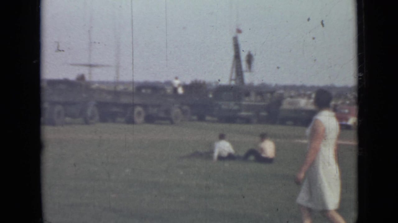Woman in a white dress walks across grass near seated people and military trucks.