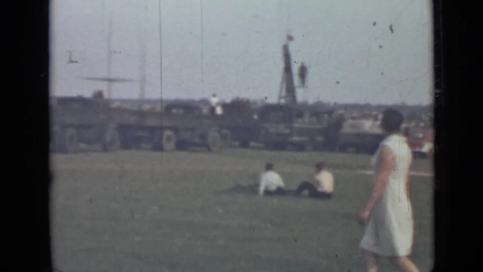 1967 Saint Petersburg home-movie frame with a woman crossing a grassy field as visitors sit near large military trucks.