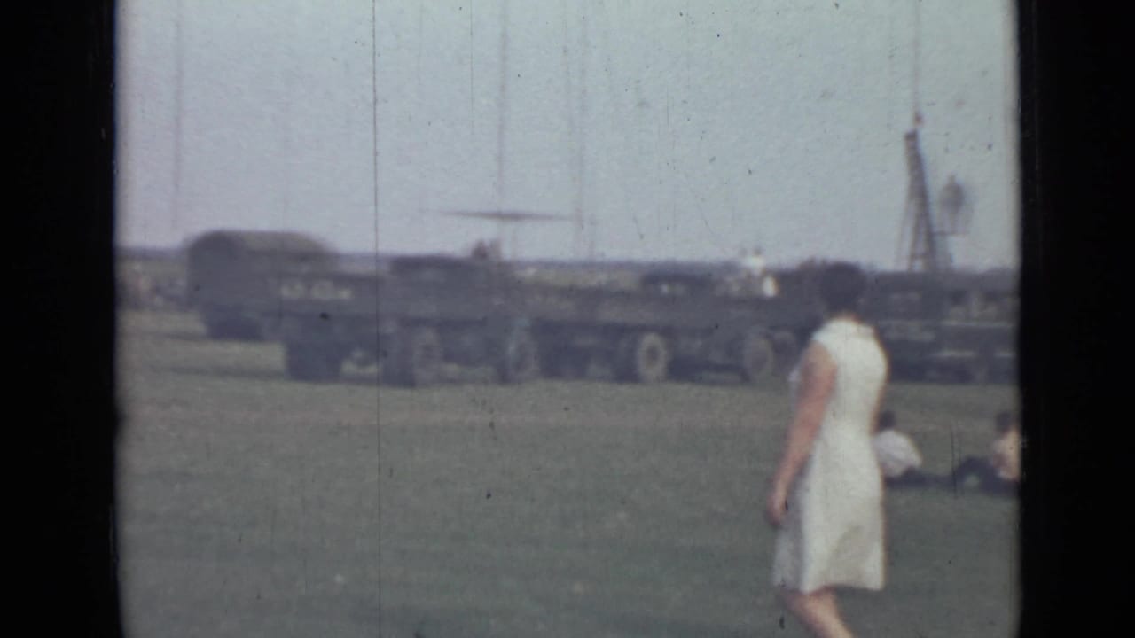 Woman in a white dress stands or walks near grass with military trucks behind her.