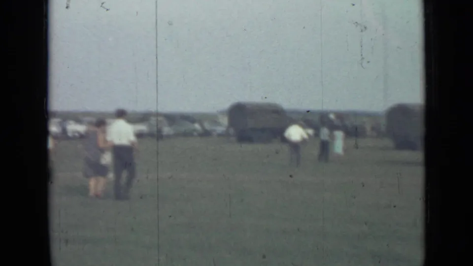 Wide 8mm frame of visitors crossing a grassy field at the 1967 Saint Petersburg military installation, with vehicles beyond them.
