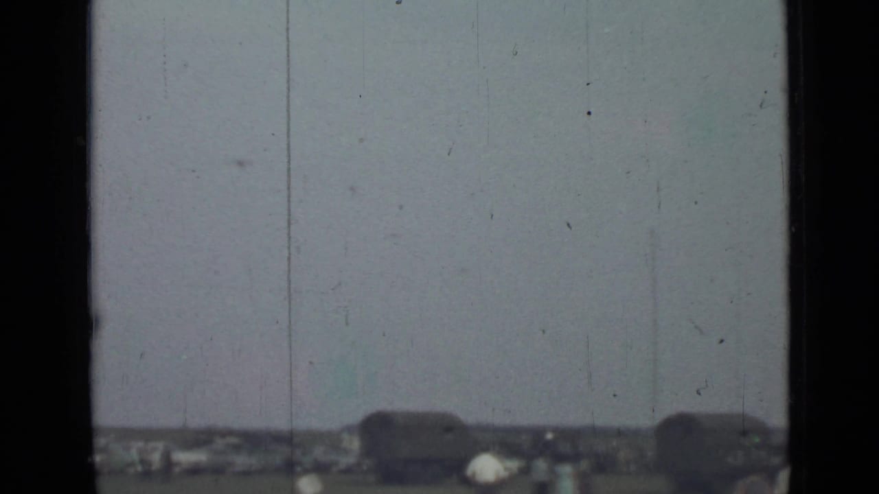 1967 Saint Petersburg home-movie frame with a low horizon, small people, dark shapes resembling vehicles or trailers, and a pale overcast sky.