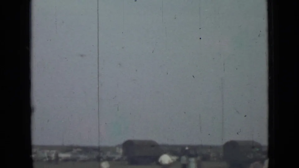 1967 Saint Petersburg home-movie frame with a low horizon, small people, dark shapes resembling vehicles or trailers, and a pale overcast sky.