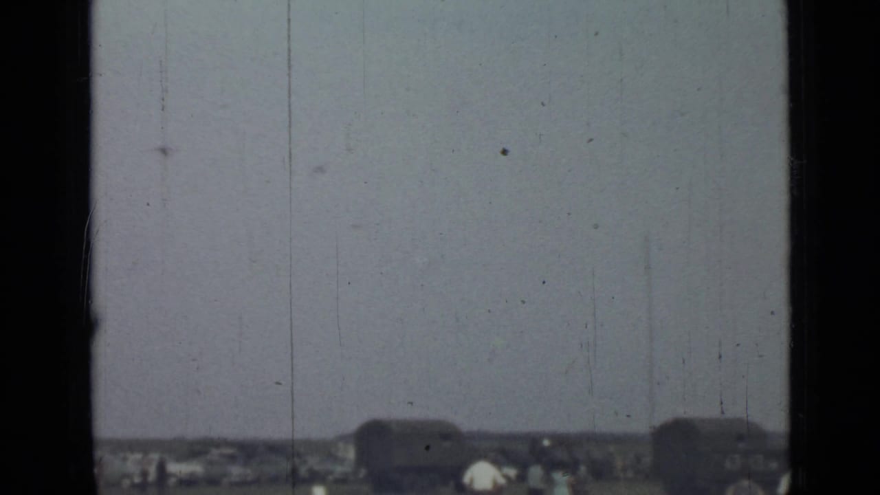 Distant 1967 Saint Petersburg exterior view with tiny figures near dark vehicle-like forms, framed by visible 8mm gate edges and scratches.