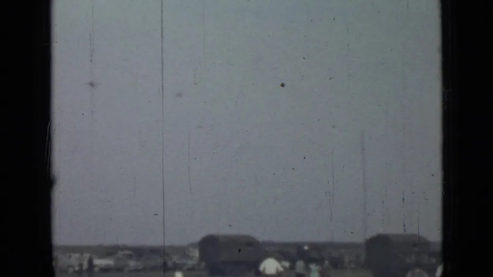 Distant 1967 Saint Petersburg exterior view with tiny figures near dark vehicle-like forms, framed by visible 8mm gate edges and scratches.