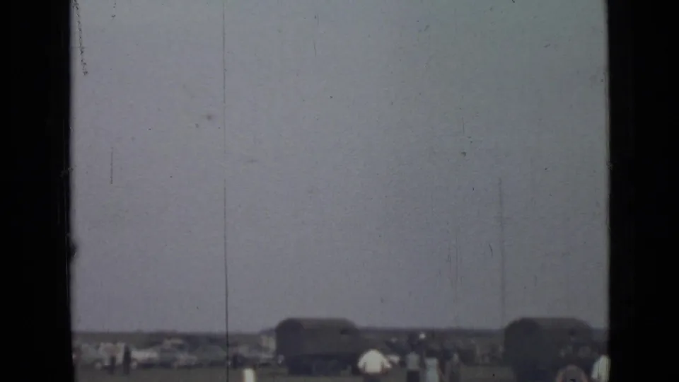 1967 Saint Petersburg 8mm frame showing a grey sky, low outdoor horizon, small people, and dark forms that may be vehicles or trailers.